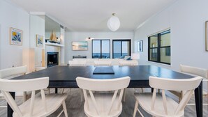 A spacious dining area with a large table and white chairs, a fireplace, and a view of the ocean through large windows. - Phoenix Ii 2151 3 Bedroom Condo (Orange Beach)