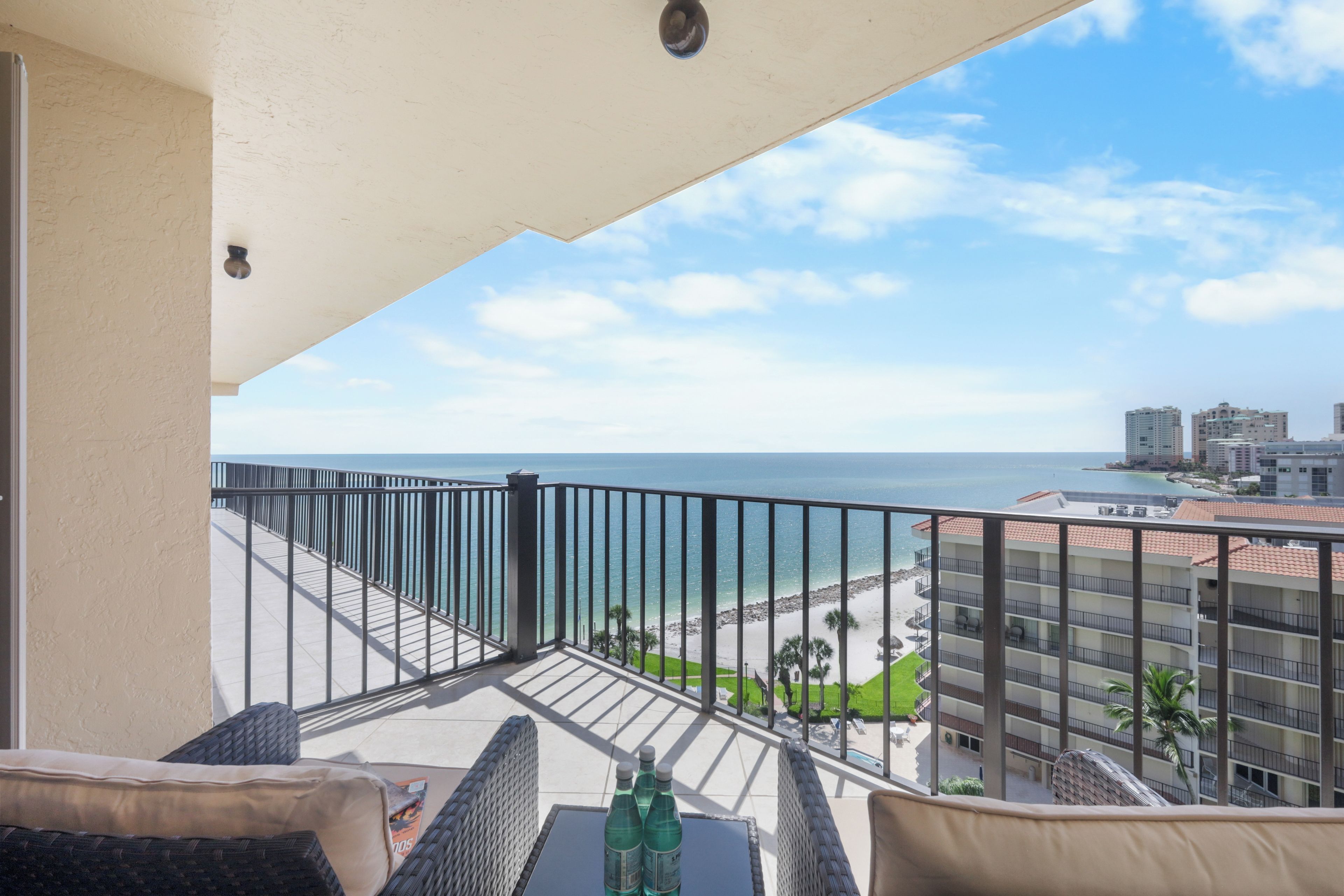 A balcony with wicker furniture, a table with bottles, and a view of the beach and buildings.
