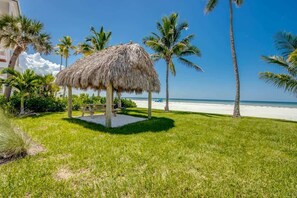 A thatched gazebo on a grassy area with palm trees and a beach in the background.