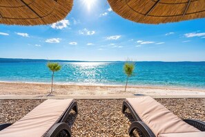 A beach view with lounge chairs, pebbles, and two small trees under a thatched roof.