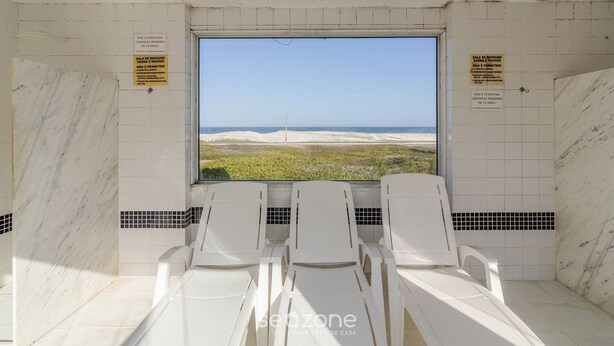 Een kamer met een groot raam met uitzicht op een strand, witte stoelen en betegelde muren.