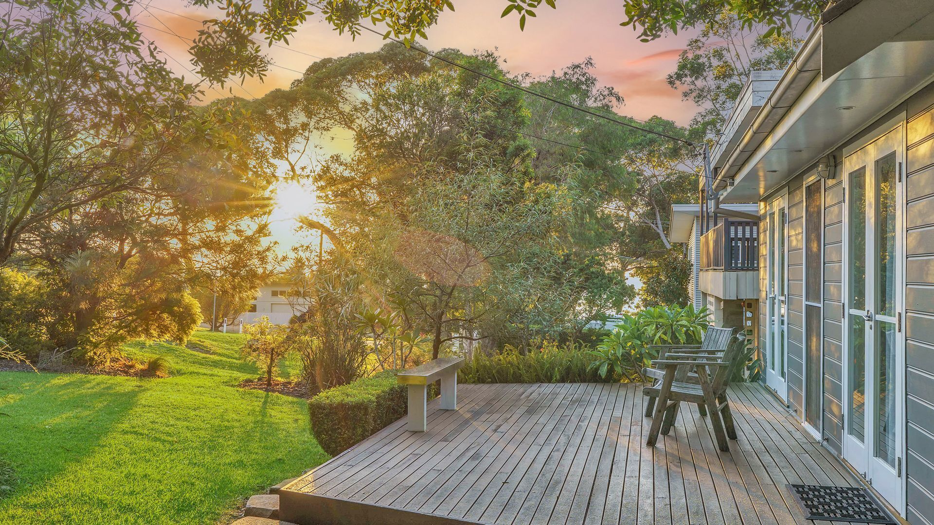 Une terrasse en bois avec un banc, entourée d’une végétation luxuriante et d’arbres, et un bâtiment doté de grandes fenêtres et d’un balcon sur la droite.