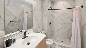 Un baño moderno con salpicadero de mármol, un lavabo blanco, un grifo negro, un inodoro y una zona de ducha con mampara de vidrio.