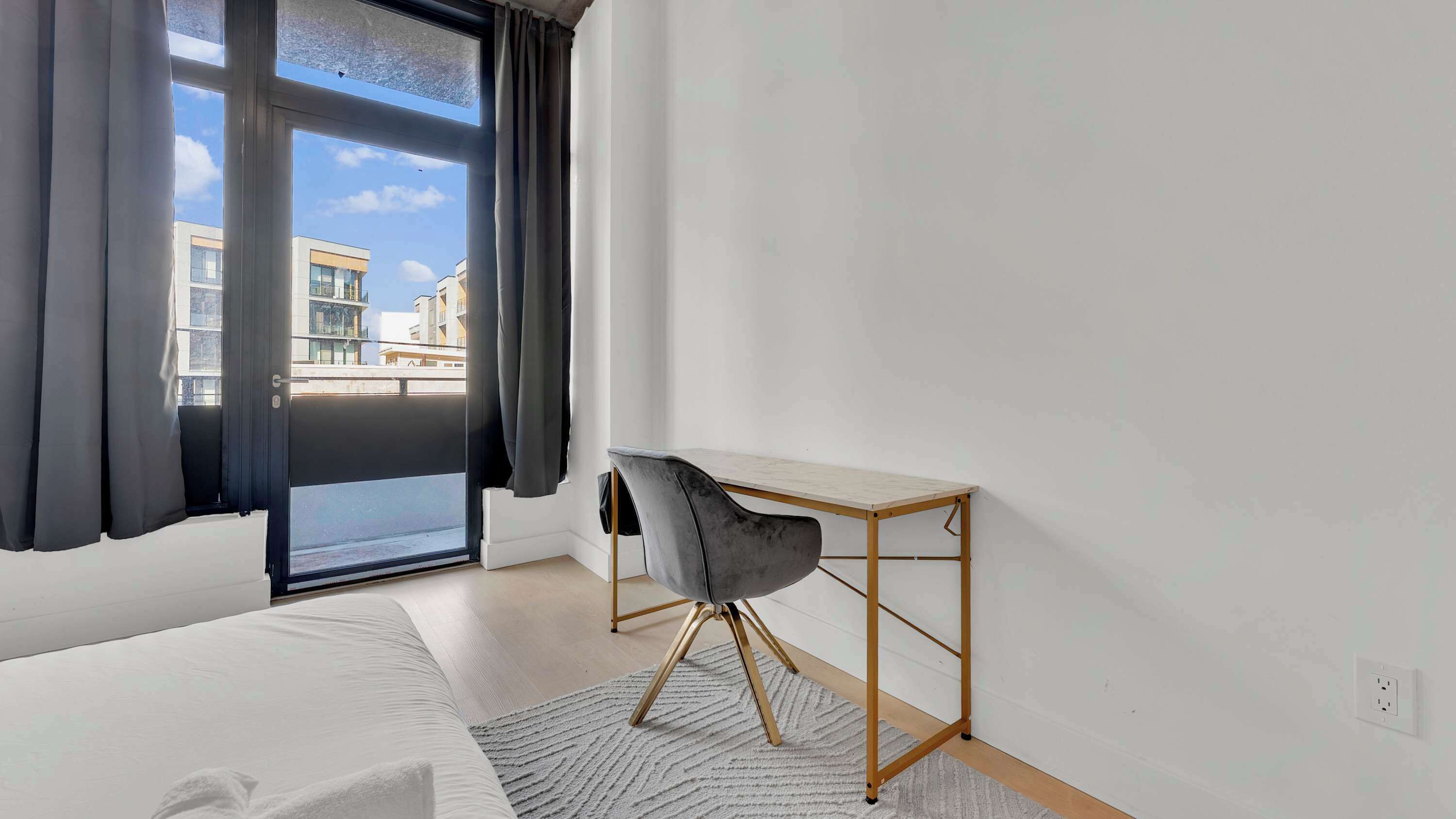 A hotel room with a bed, a desk, and a view of buildings through a window.