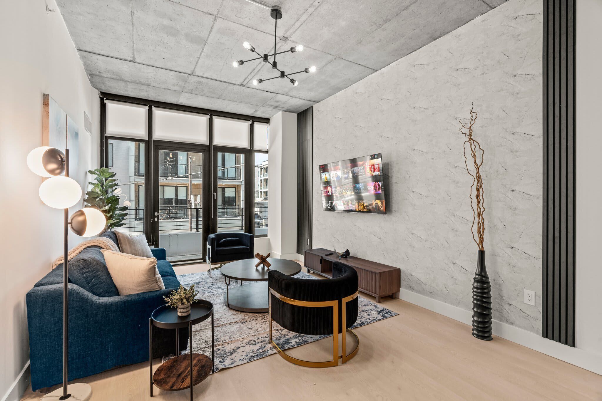 Ein modernes Wohnzimmer mit einem blauen Sofa, einem runden Couchtisch und einem schwarzen Akzentstuhl. Es gibt eine Stehlampe, einen kleinen Tisch mit einer Pflanze und eine hohe Vase mit Zweigen.