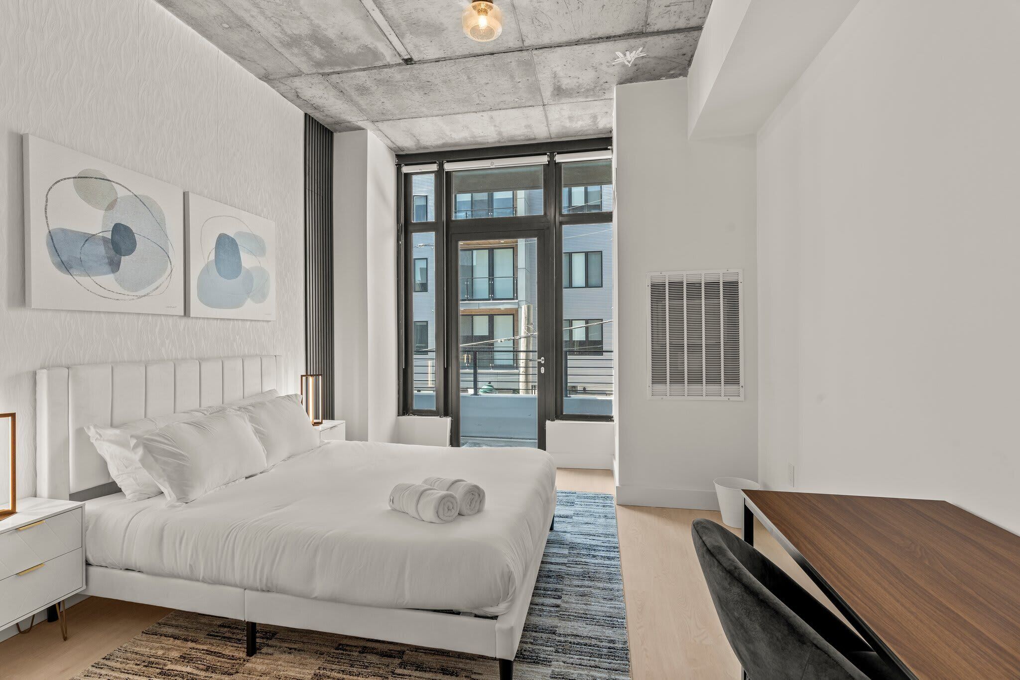 Ein modernes Schlafzimmer mit einem großen Bett, einem Holztisch und Blick auf Gebäude durch große Fenster.