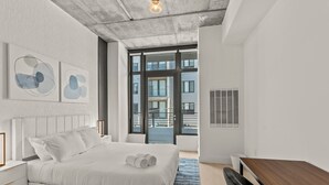 Un dormitorio moderno con una cama grande, un escritorio de madera y vistas a edificios a través de amplios ventanales.