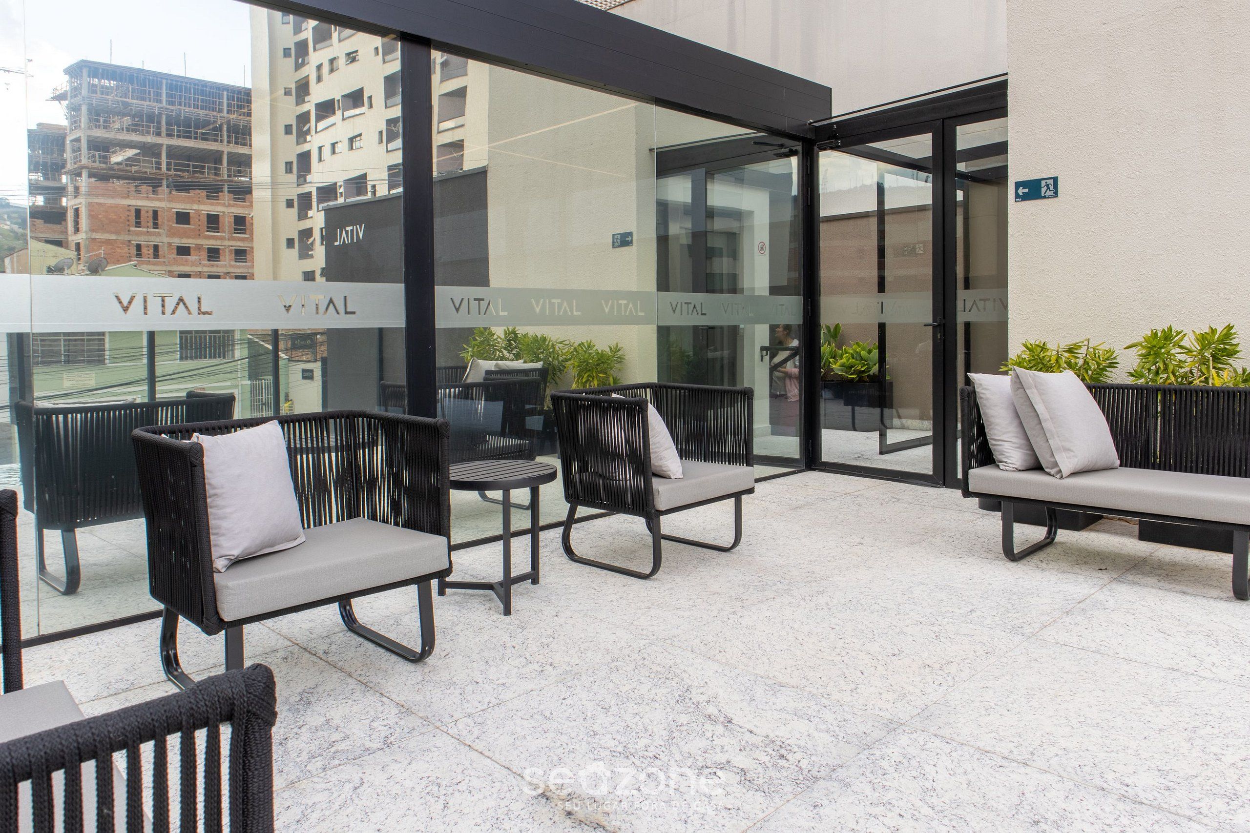 A modern outdoor seating area with wicker chairs and a bench, featuring a glass wall with 'VITAL' signage, potted plants, and a cityscape view.
