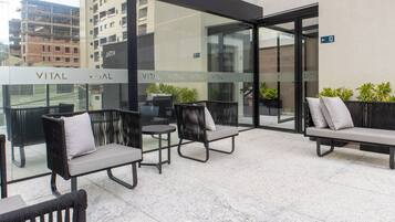 A modern outdoor seating area with wicker chairs and a bench, featuring a glass wall with 'VITAL' signage, potted plants, and a cityscape view.