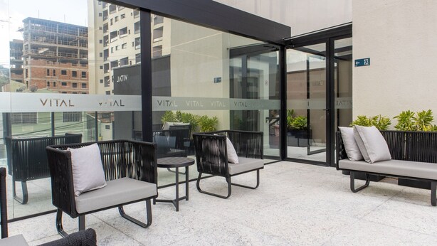 A modern outdoor seating area with wicker chairs and a bench, featuring a glass wall with 'VITAL' signage, potted plants, and a cityscape view. - Your Complete Stay With Pool and gym in Pocos de Caldas - NGO (Poços de Caldas)