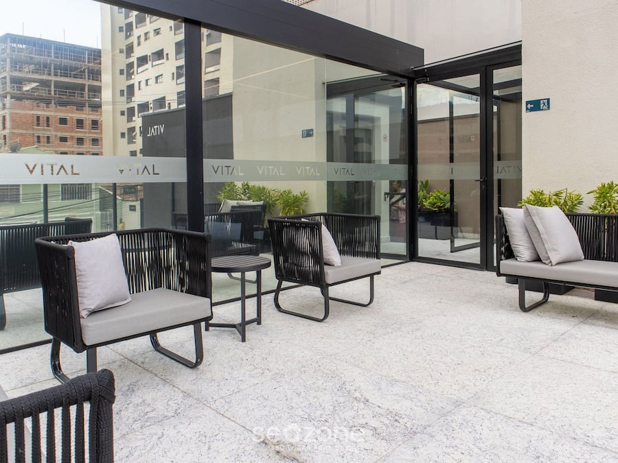Un área de estar moderna al aire libre con sillas de mimbre y un banco, con un muro de cristal que muestra el letrero 'VITAL', plantas en macetas y vistas de la ciudad.