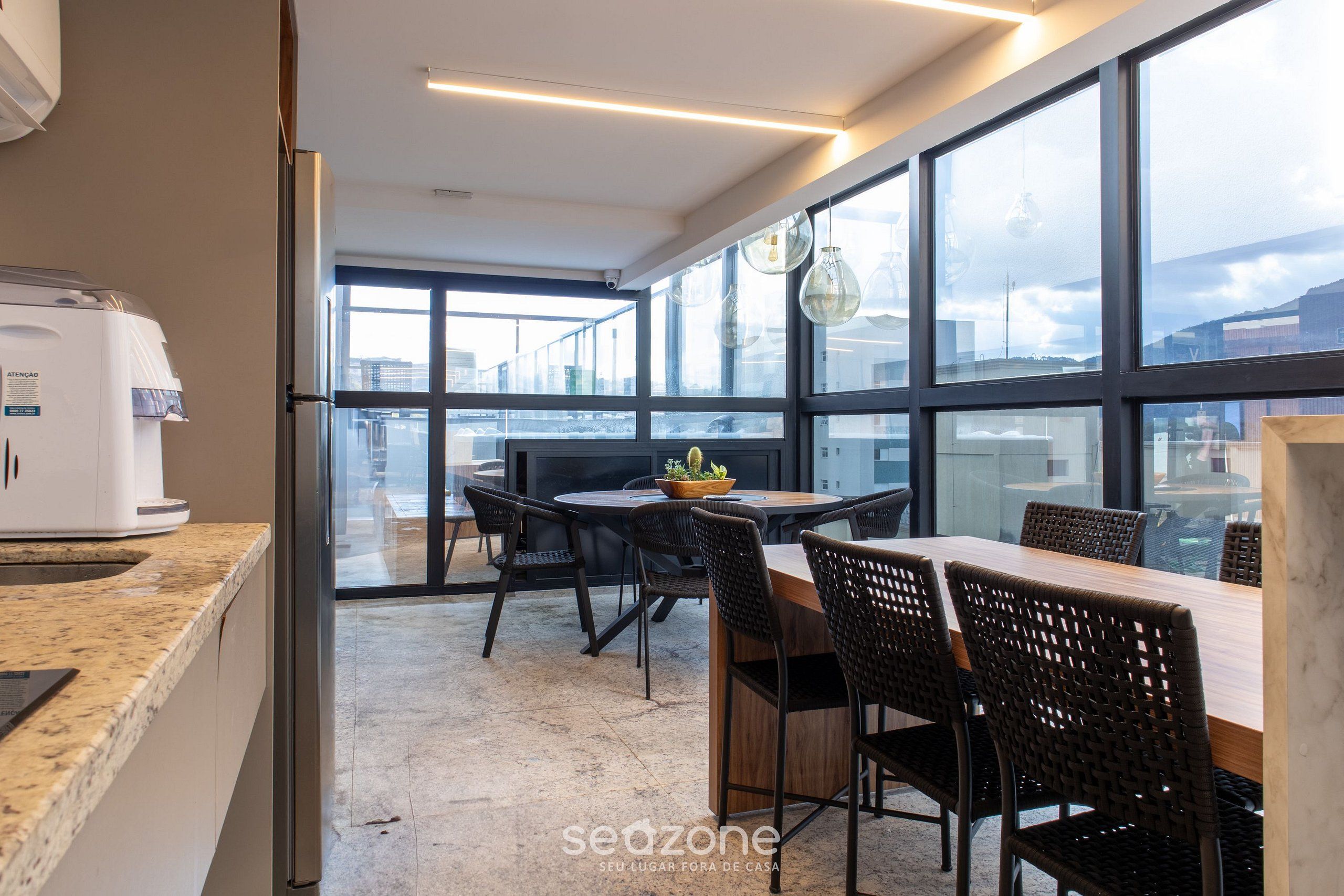 A modern kitchen with a coffee machine, a dining area with a table and chairs, and large windows offering a view of the city.