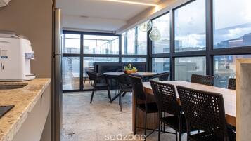 A modern kitchen with a coffee machine, a dining area with a table and chairs, and large windows offering a view of the city.