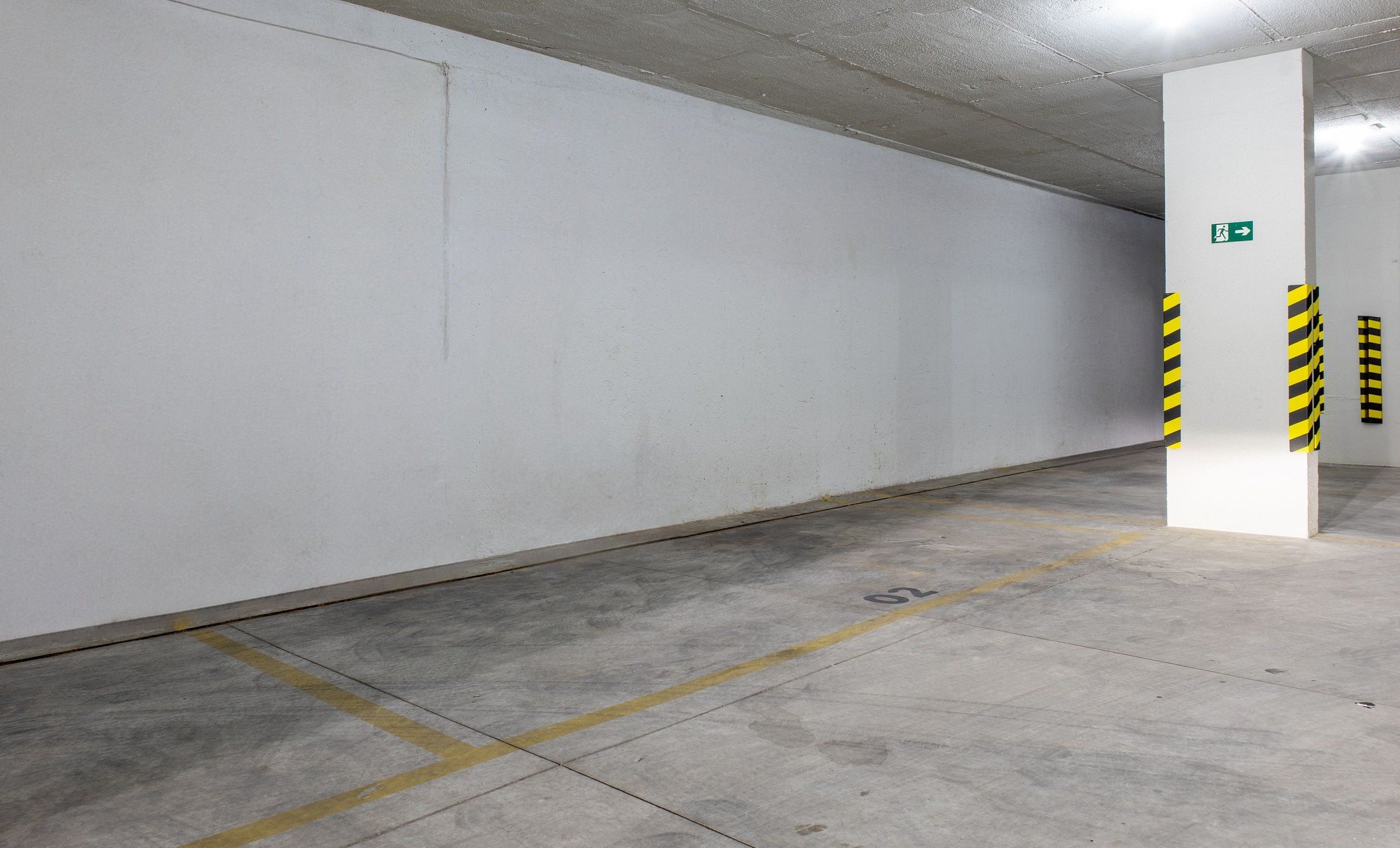 Un parking propre et vide, avec des murs blancs, des lignes de stationnement jaunes et un panneau vert indiquant la sortie de secours.