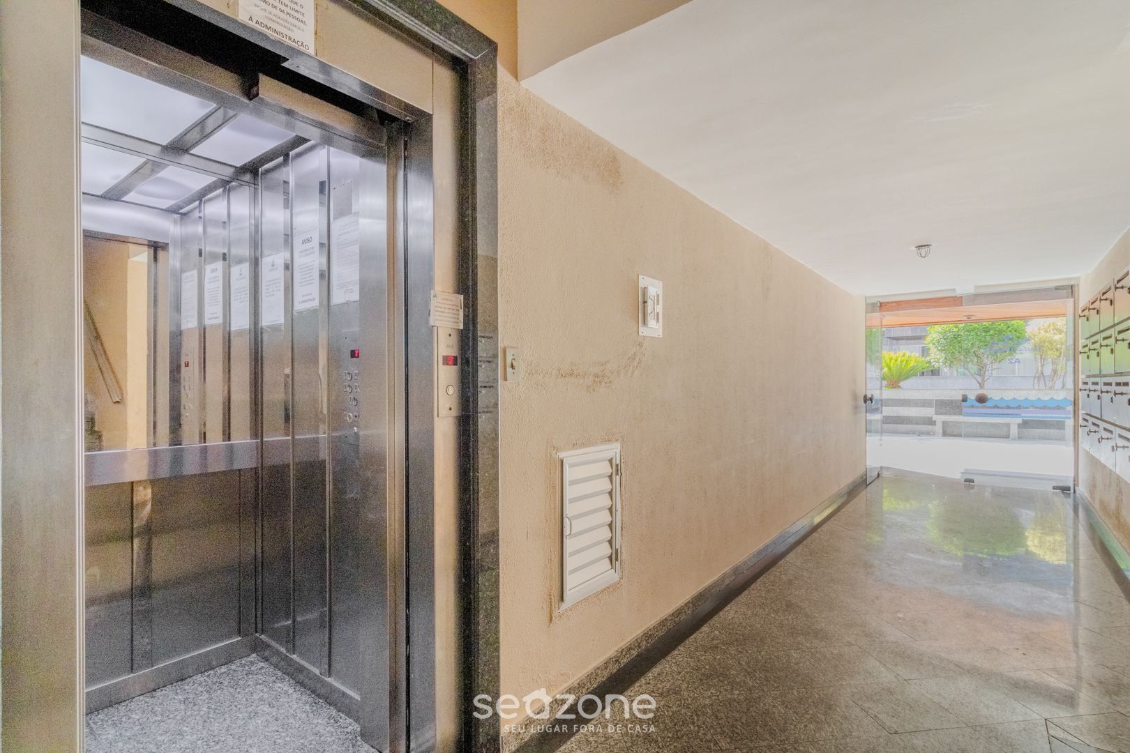 An elevator in a hallway with a beige wall, a small window, and a door leading to a room.