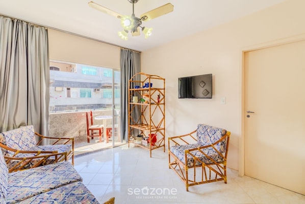A room with a balcony, two wicker chairs, a wooden shelf, a ceiling fan, and a TV on the wall.