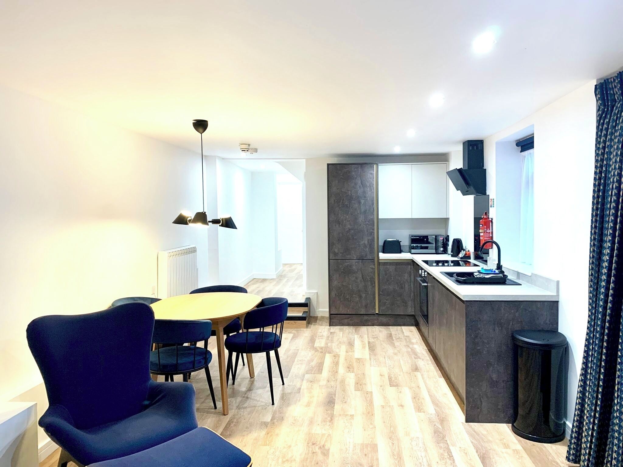 A modern kitchen with a built-in oven, microwave, and coffee machine. There is a dining area with a round table and blue chairs. The room has a wooden floor and a large window with curtains.