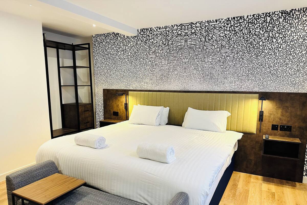 Quarto de hotel moderno com uma cama grande, duas mesas de cabeceira, uma pequena mesa de madeira, uma poltrona cinzenta e uma parede estampada.