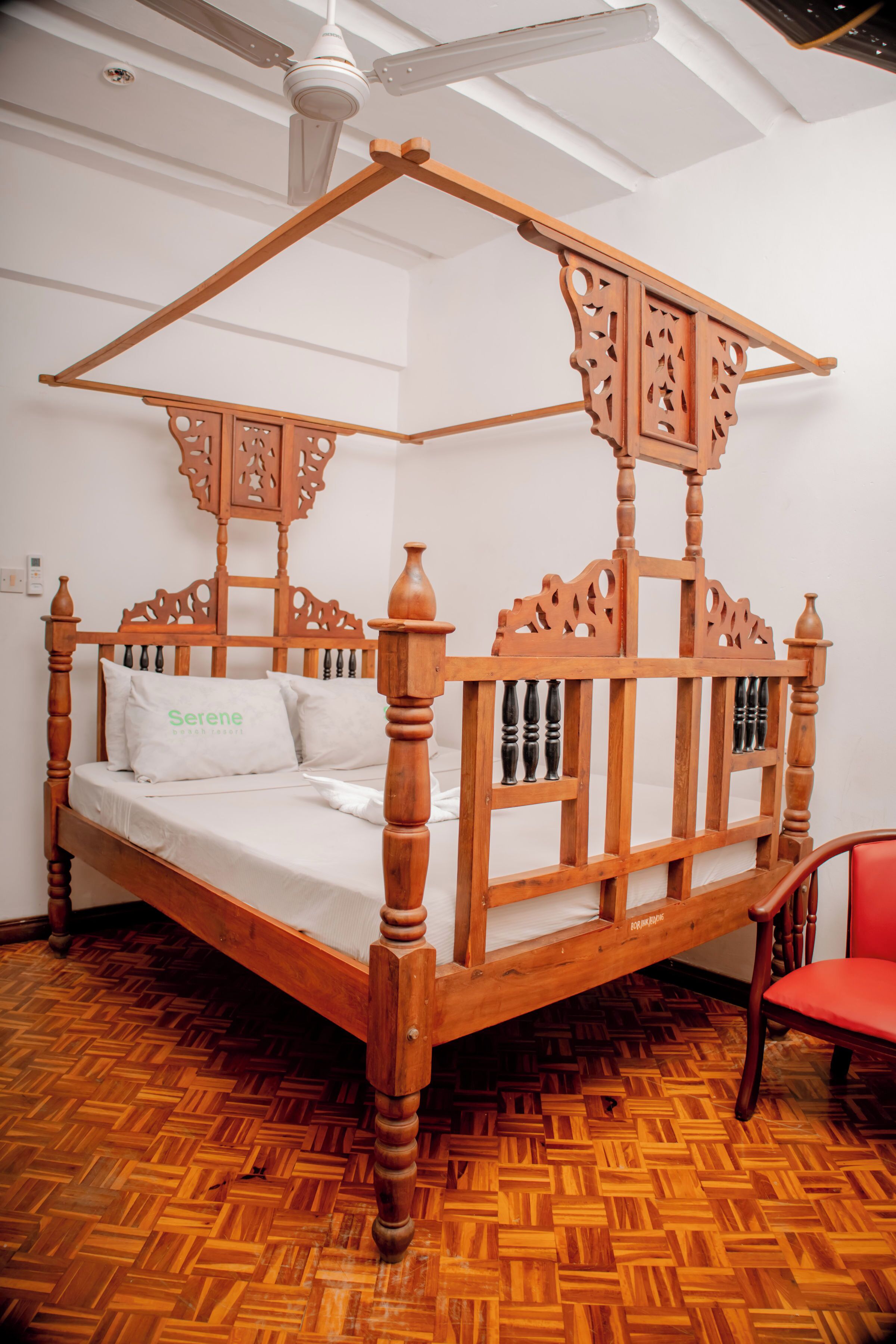 A wooden four-poster bed with a white mattress and pillows, a red chair, and a herringbone-patterned wooden floor.