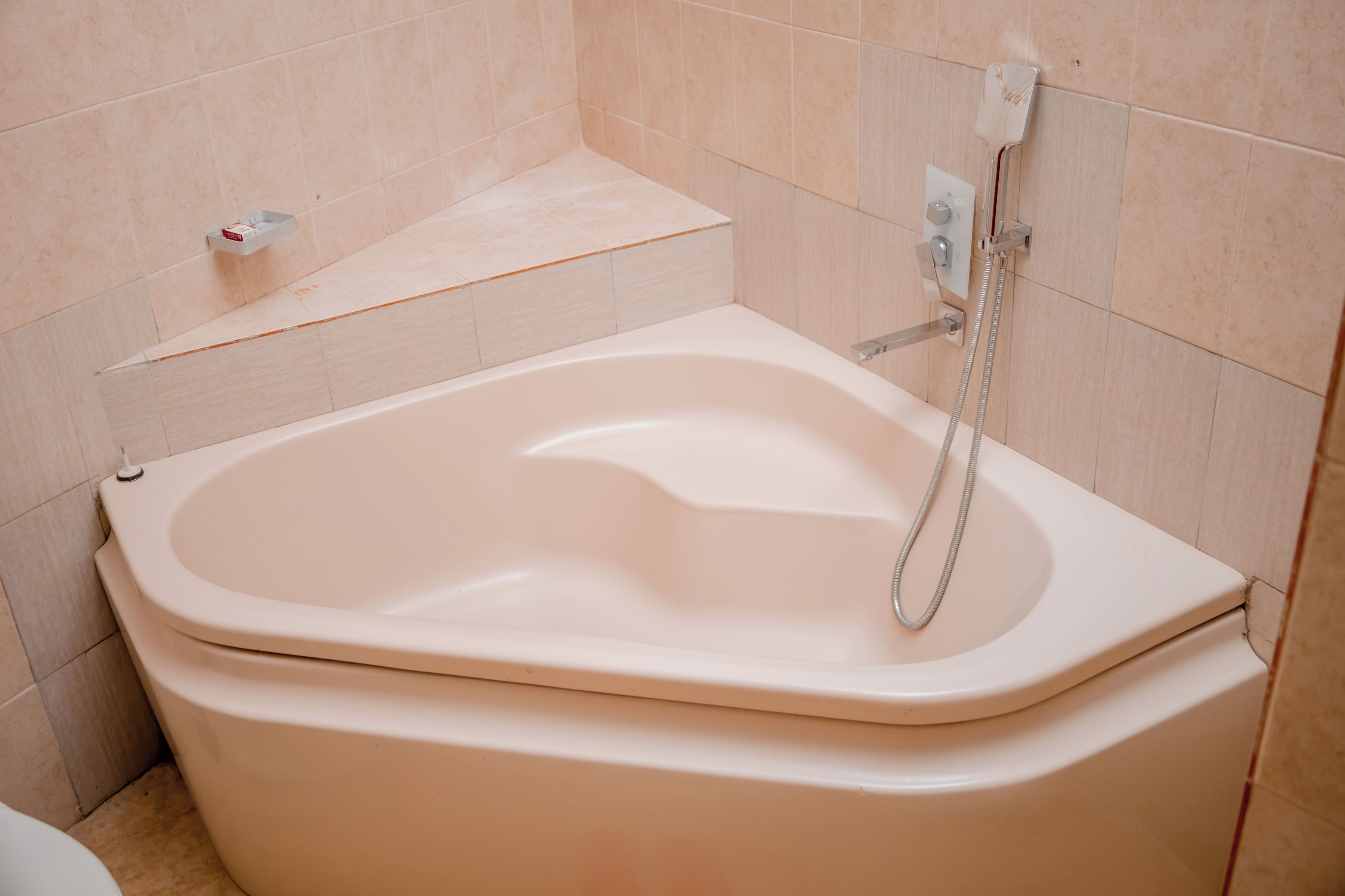 A standard bathtub with a handheld showerhead in a bathroom with beige tiles.