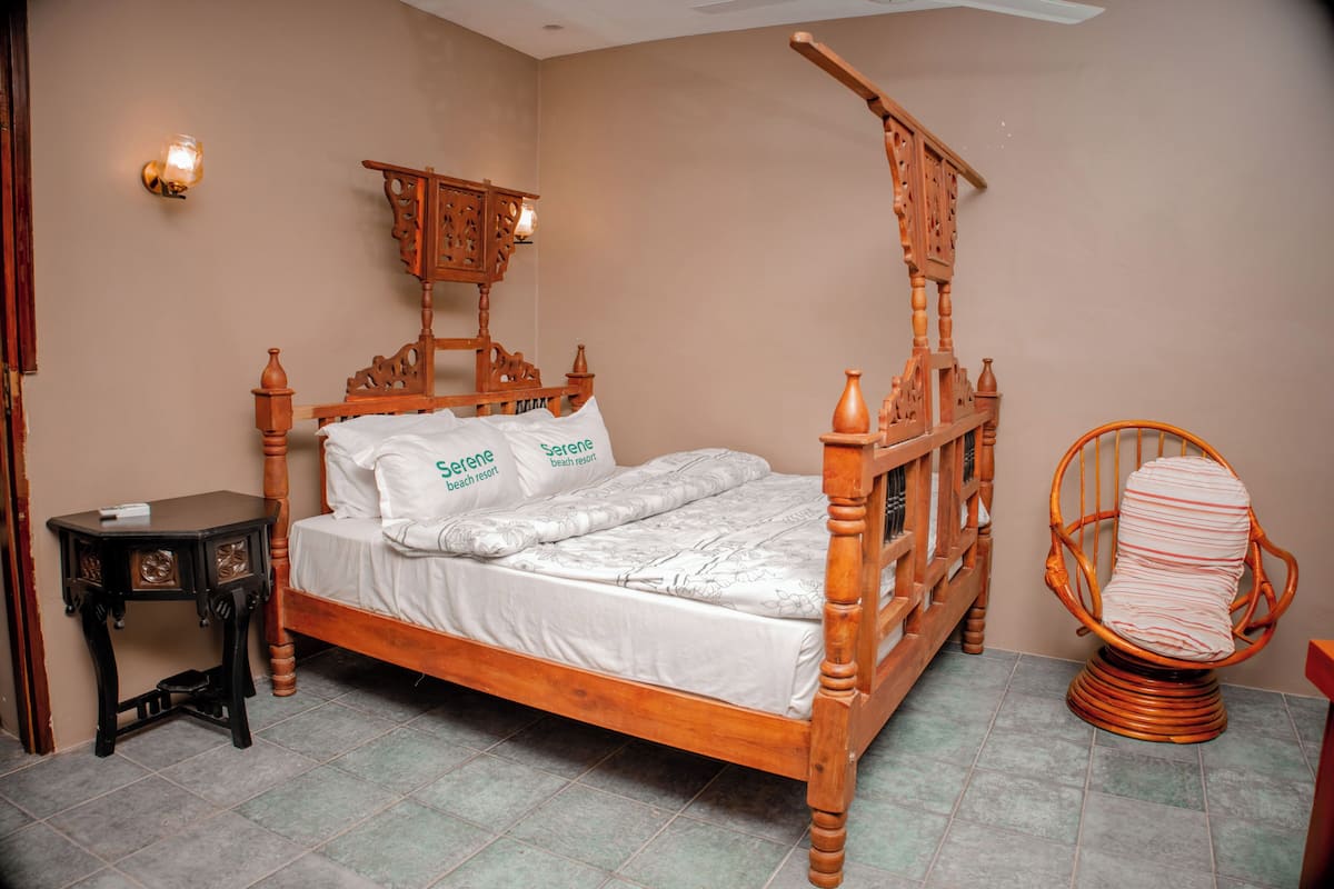 A wooden four-poster bed with a white mattress and pillows, a small bedside table, and a wicker chair.