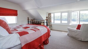 Un dormitorio con cama, un sillón blanco, un ventanal con vista a un lago y un edredón rojo y blanco.