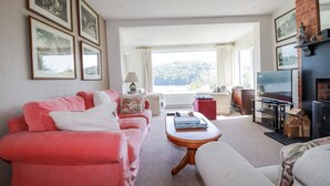 Una sala de estar con un sofá rojo, un sofá blanco, una mesa de centro de madera y una chimenea. Hay cuadros en la pared y un televisor sobre un mueble.