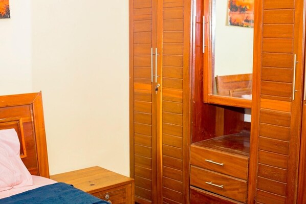 A bedroom with a wooden wardrobe, a bed with a pink pillow, and a small bedside table.