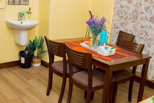 Un comedor con una mesa de madera, cuatro sillas, un florero con flores moradas, una jarra de agua azul y una bandeja con condimentos.
