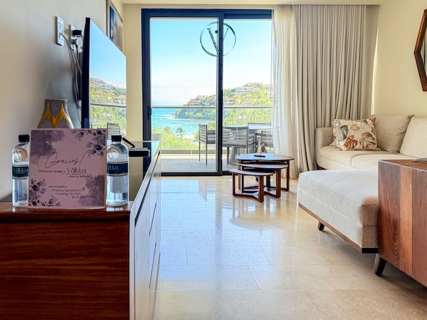 A hotel room with a balcony, a sofa, a coffee table, and a desk with a sign and water bottles.