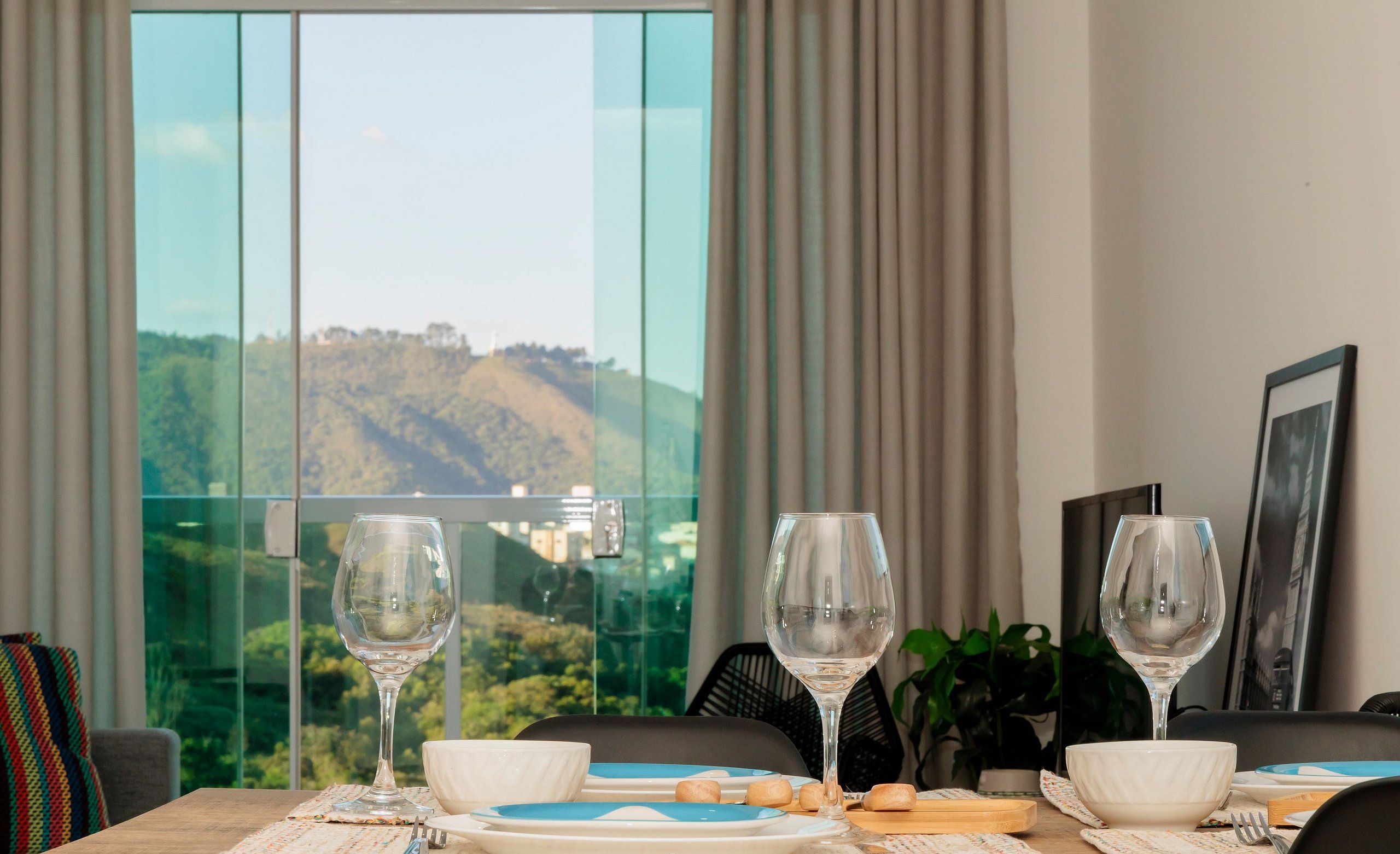 Una mesa de comedor con platos, tazones y copas de vino, con vistas panorámicas de colinas a través de amplios ventanales.
