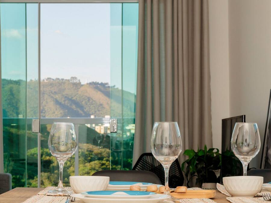 A dining table set with plates, bowls, and wine glasses, with a scenic view of hills through large windows.