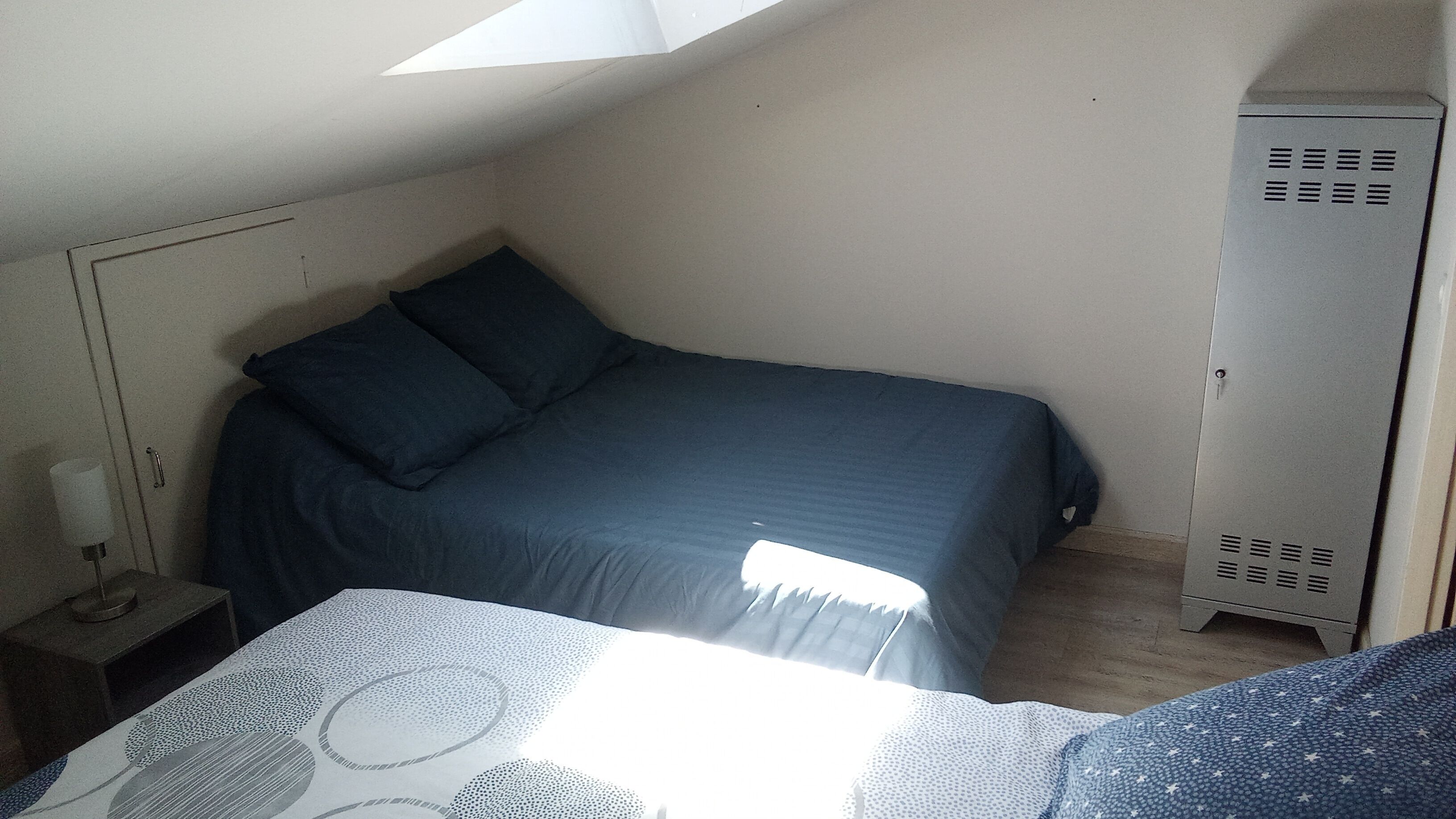 Ein kleiner Raum mit gedämpftem Licht, einem Bett, einem Nachttisch, einem Kleiderschrank und einer schrägen Decke.