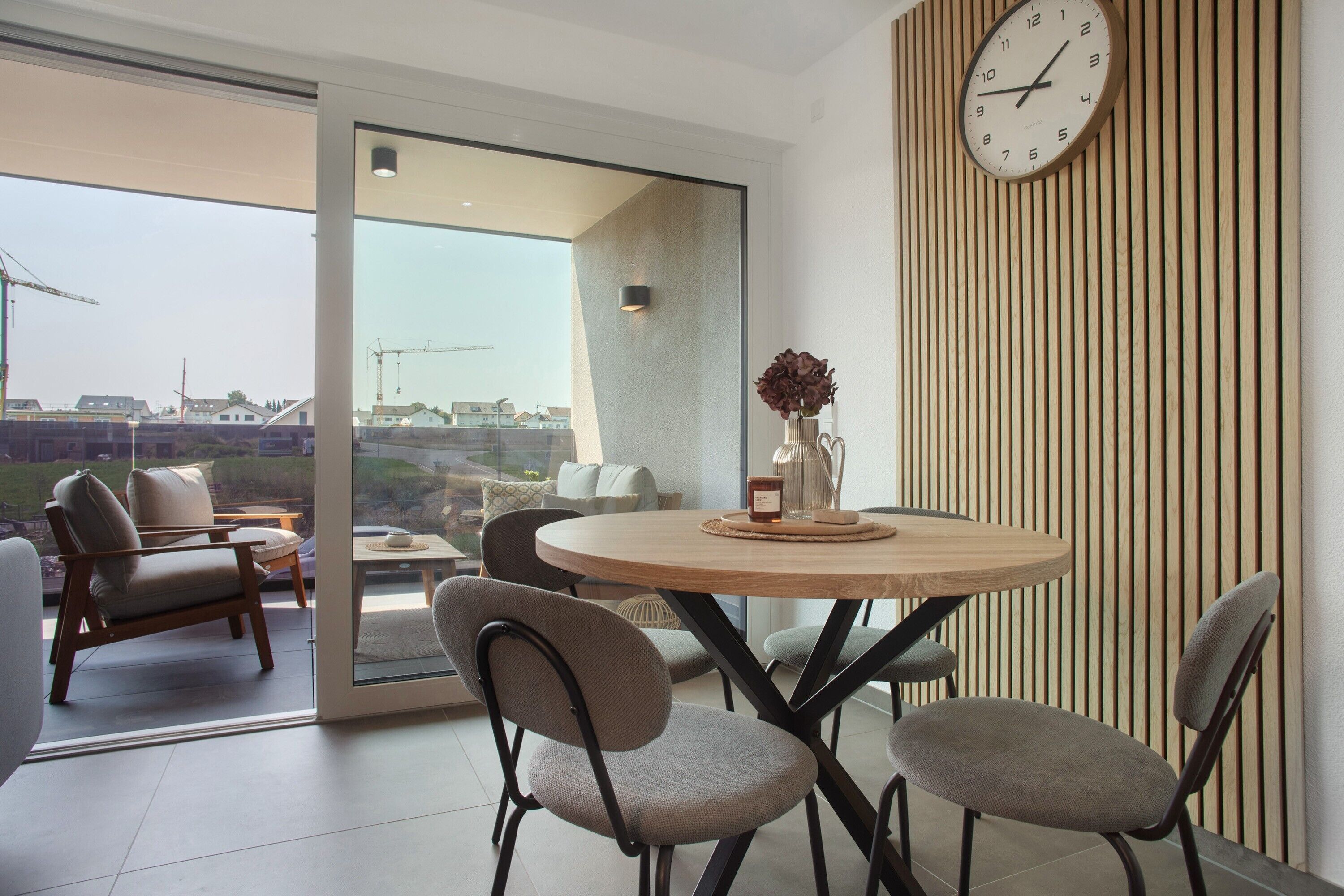 A modern dining area with a round table, four chairs, a clock, and a view of construction cranes outside.