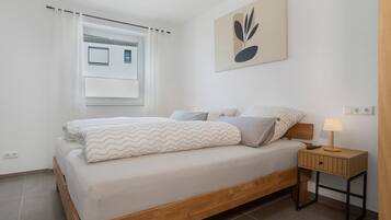 A modern bedroom with a wooden bed, a bedside table with a lamp, a window with curtains, and a wall-mounted abstract art piece.