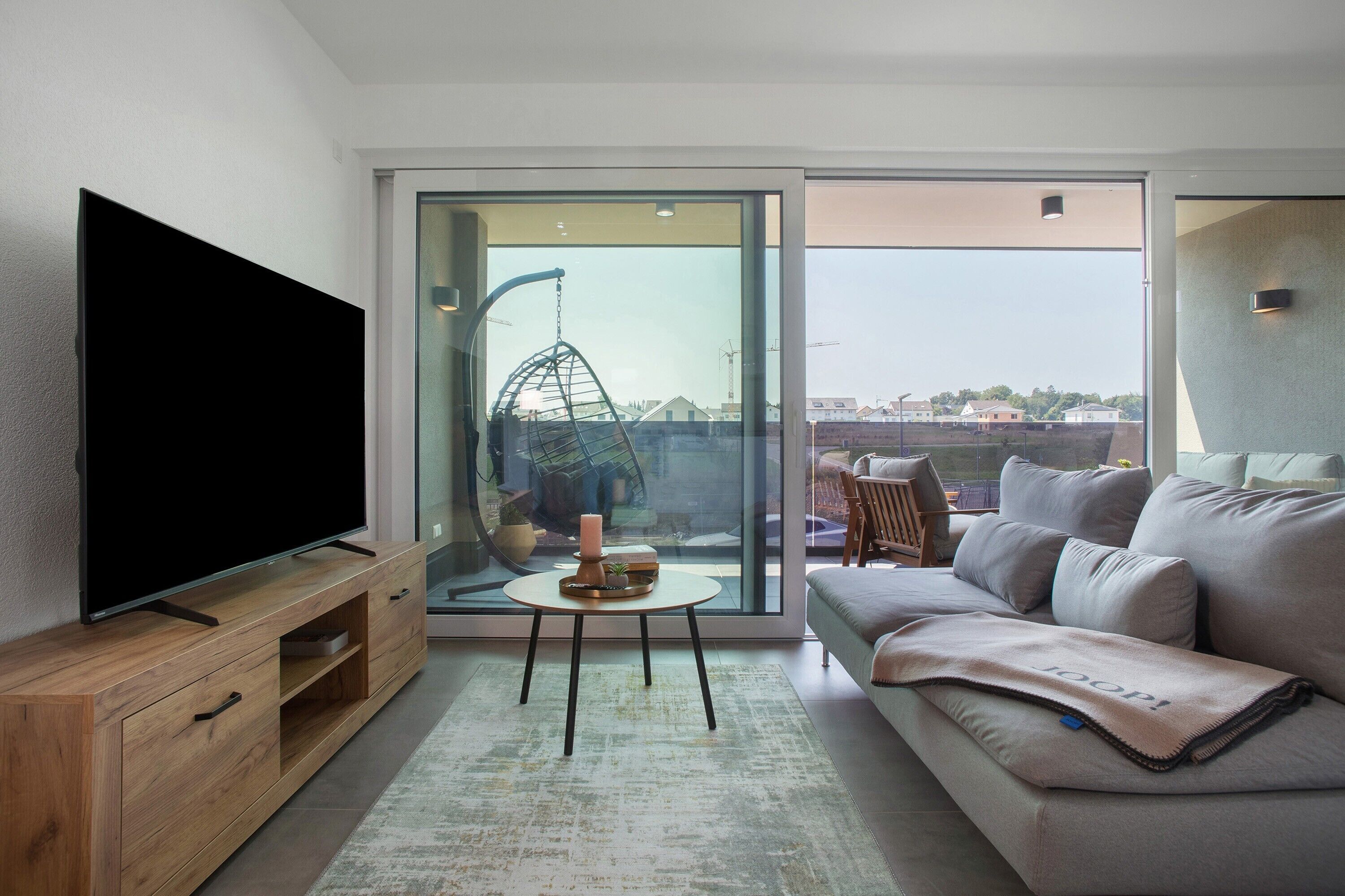 Un salón moderno con televisor de pantalla plana, un sofá, una mesita con una vela y vistas al exterior a través de un ventanal.