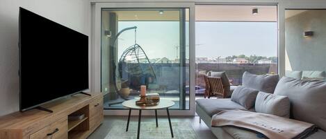 Un salón moderno con televisor de pantalla plana, un sofá, una mesita con una vela y vistas al exterior a través de un ventanal.