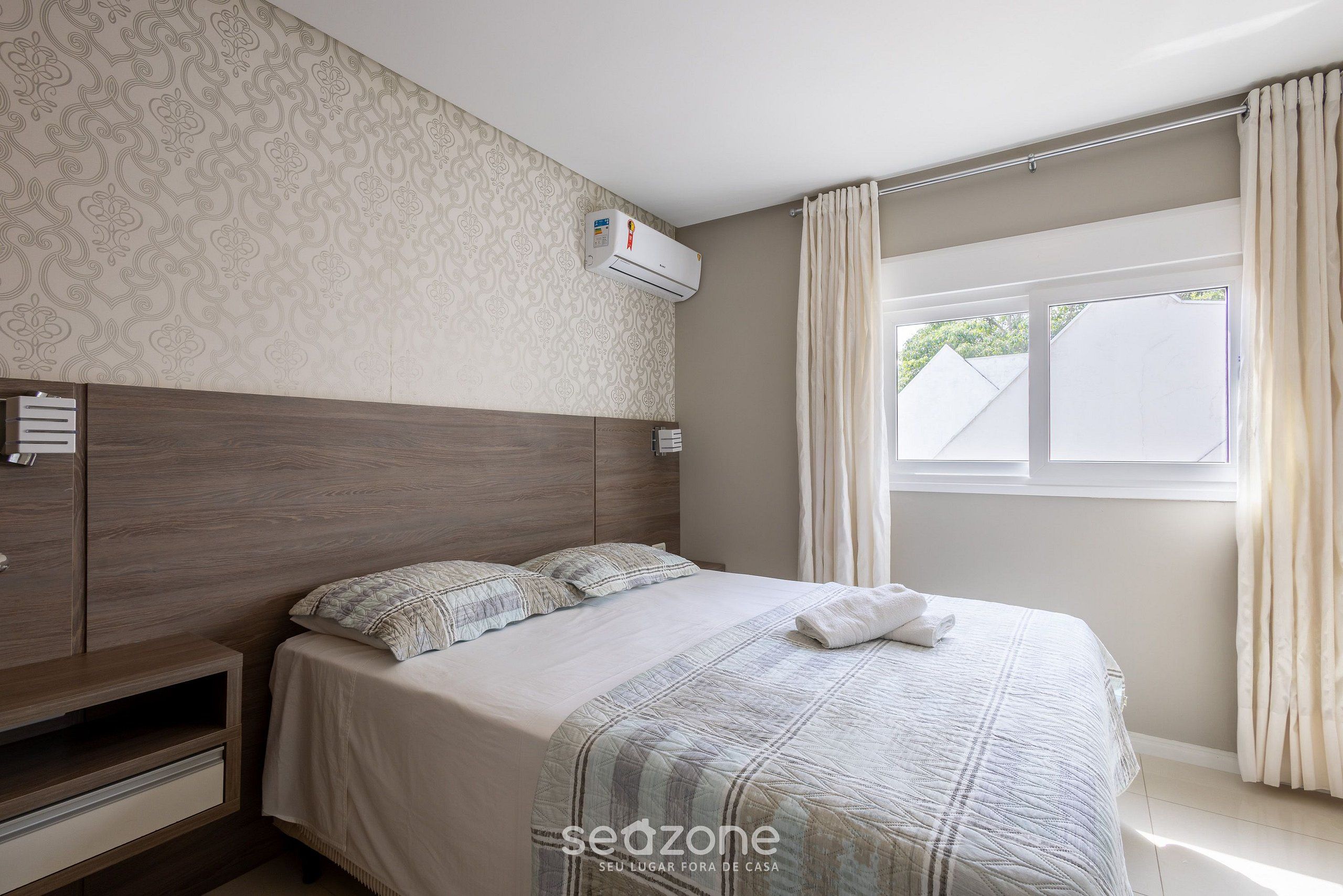 A bedroom with a wooden headboard, a bed with a striped blanket, a bedside table, and a window with curtains.
