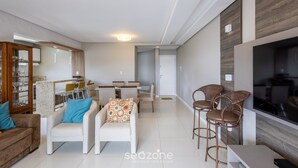 A modern living room with a sofa, armchairs, a dining area, and a TV. - Sea View Apartment w Barbecue in Bombinhas Lga0201 (Bombinhas)
