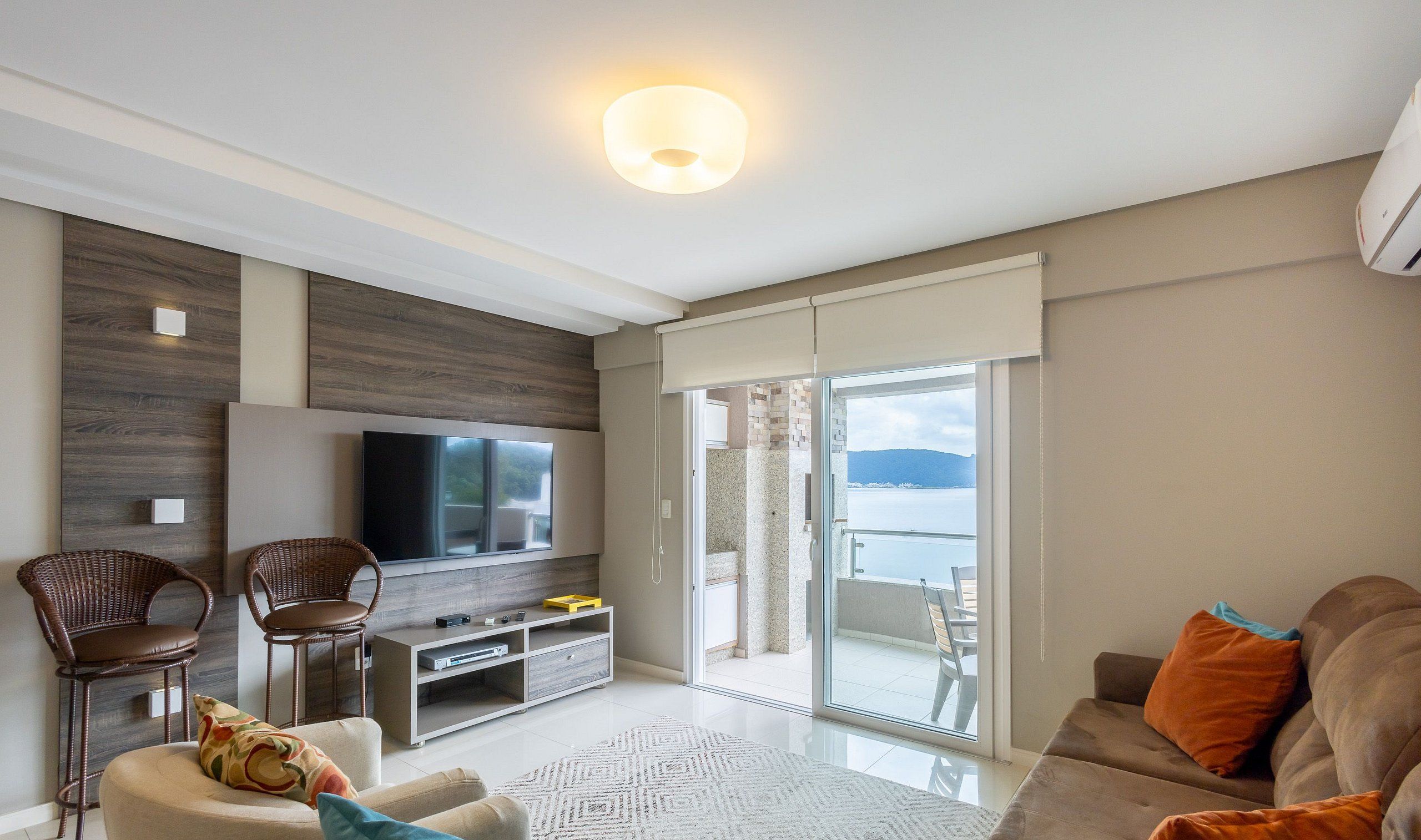 A modern living room with a flat-screen TV, wicker chairs, a sofa, and a balcony with a view of the sea.