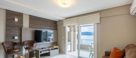 A modern living room with a flat-screen TV, wicker chairs, a sofa, and a balcony with a view of the sea.
