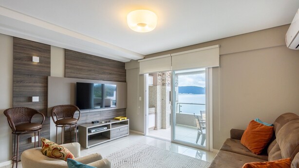 A modern living room with a flat-screen TV, wicker chairs, a sofa, and a balcony with a view of the sea. - Sea View Apartment w Barbecue in Bombinhas Lga0201 (Bombinhas)