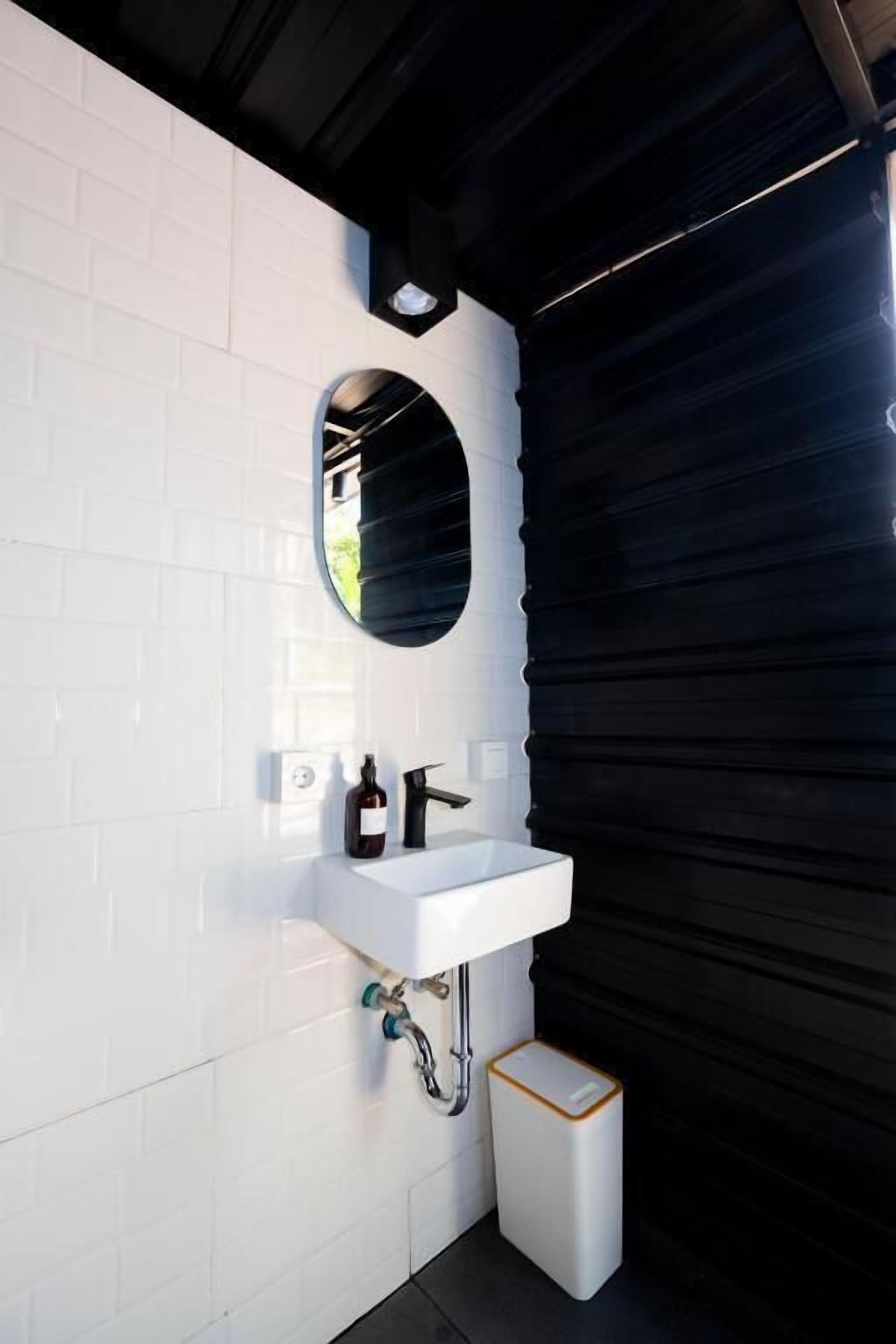 A modern bathroom with a white tiled wall, a white sink, a black faucet, a mirror, and a white trash bin.