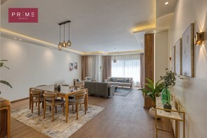A modern living room with a dining area, a sofa set, and a large window with curtains. - Prime Select Village Gardens Kattameya (New Cairo)