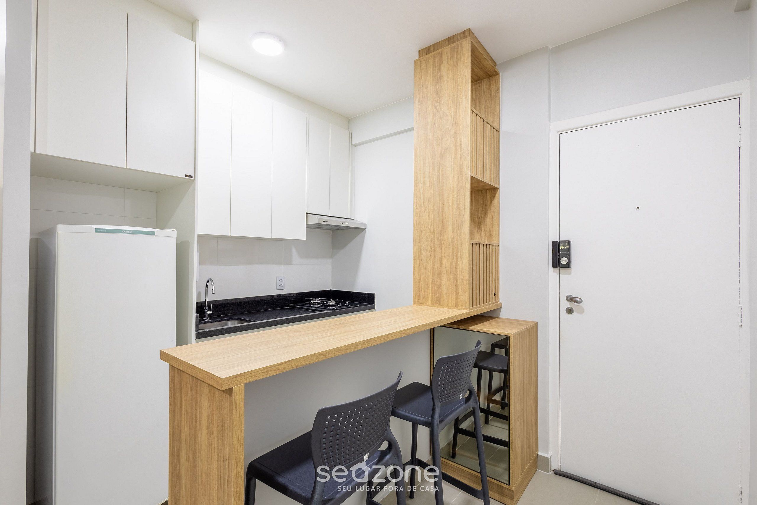 A compact kitchen with white cabinets, a wooden countertop, and a built-in stove. There are two black chairs and a small wooden shelf.
