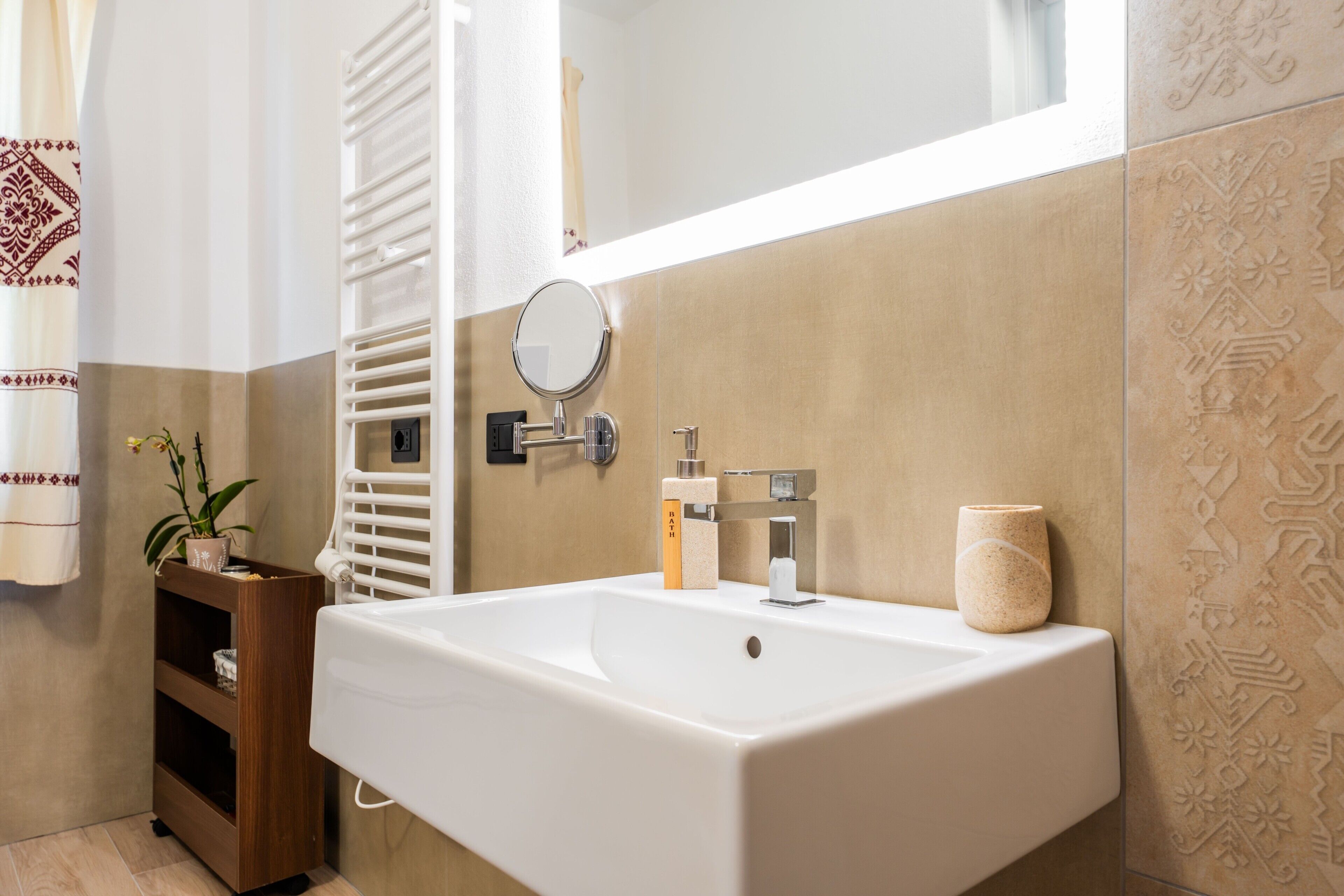A bathroom with a white sink, a mirror, a towel rack, a shelf with a plant, and patterned tiles.