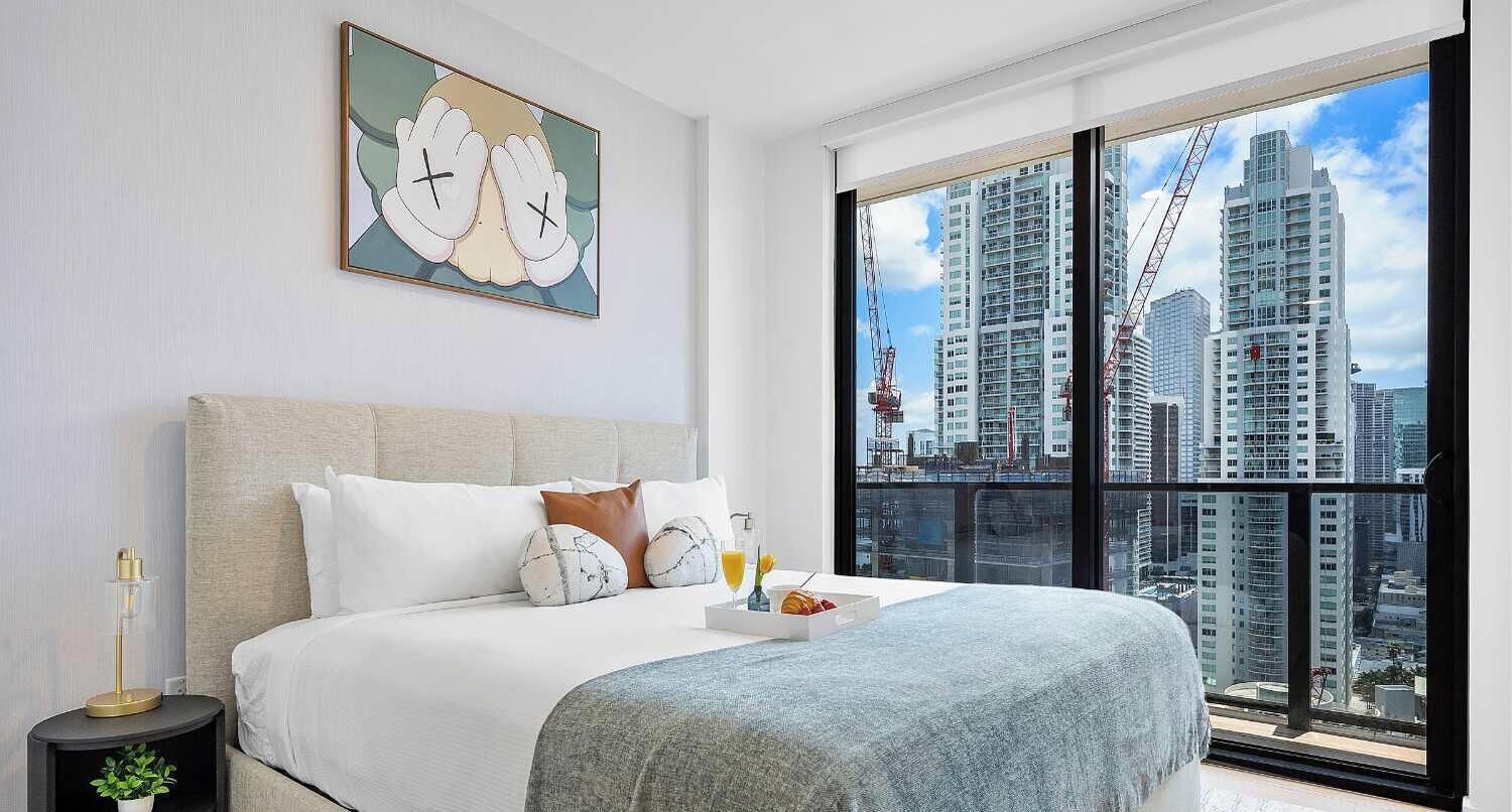 A modern hotel room with a large bed, a city view, and a painting on the wall.