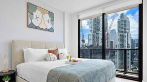 A modern hotel room with a large bed, a city view, and a framed artwork on the wall.