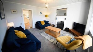 A living room with a sofa, armchair, coffee table, TV, fireplace, and a door leading to a kitchen. - The perfect base for exploring the island with your family. (Portree)