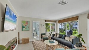 A spacious living room with a large sofa, a coffee table, and a television mounted on the wall. There is a window with a view of greenery outside.